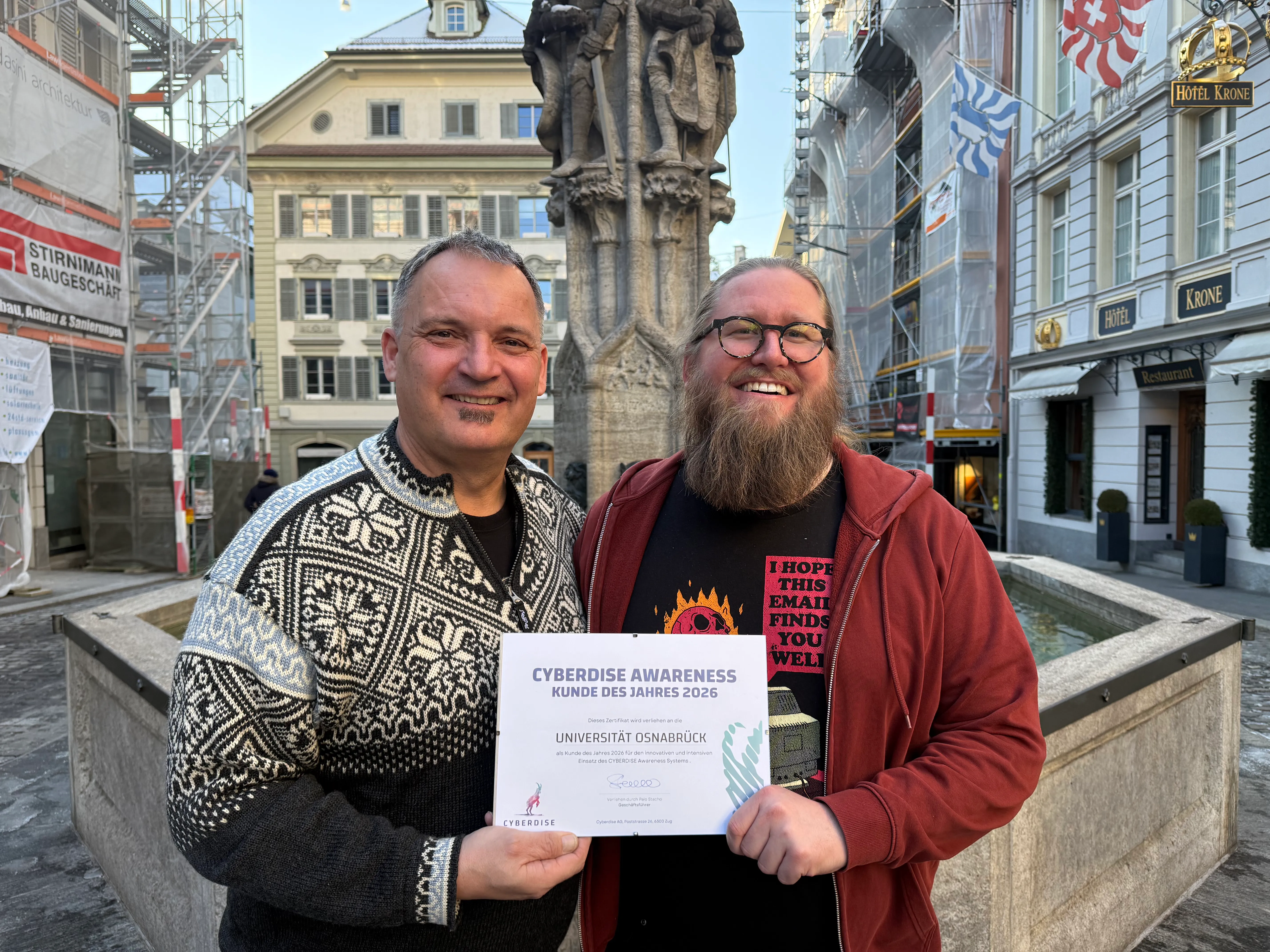 Two men at an old-town fountain hold the "Cyberdise Awareness Kunde des Jahres 2026" certificate for Universität Osnabrück