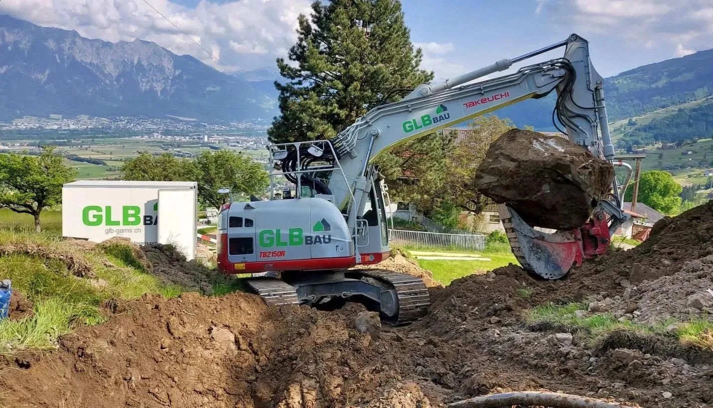 Ein Bagger von GLB Bau auf einer Baustelle am Hang, mit Bergen im Hintergrund