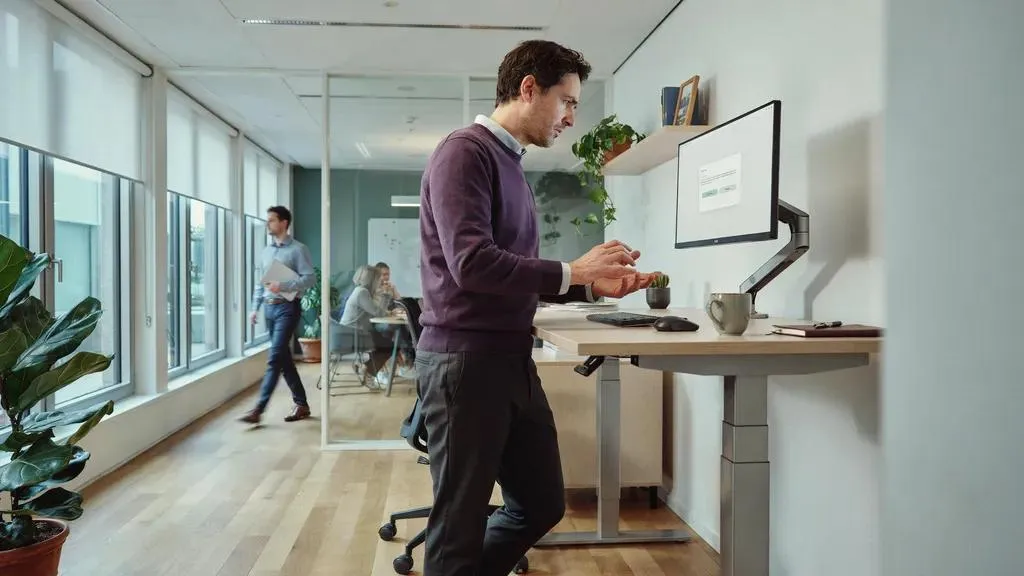 A man at a standing desk in a bright office pauses with his hands lifted away from the keyboard, hesitating in front of a dialog box on his monitor